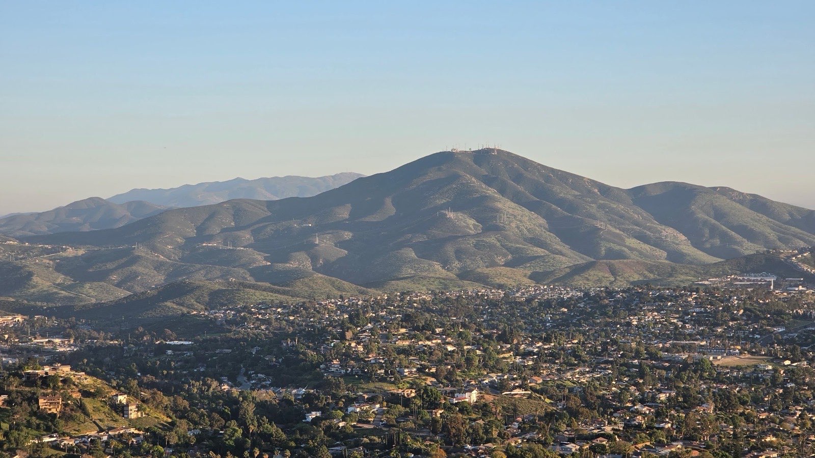 A view across La Mesa at golden hour. Houses and trees fill the valley below, and a broad hill topped with radio towers rises behind it.